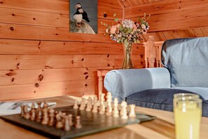 A wooden cabin interior with a chessboard, a sofa, a vase with flowers, and a framed picture of a puffin. - 3 Bed in Little Petherick (oc-c28311) (Little Petherick)