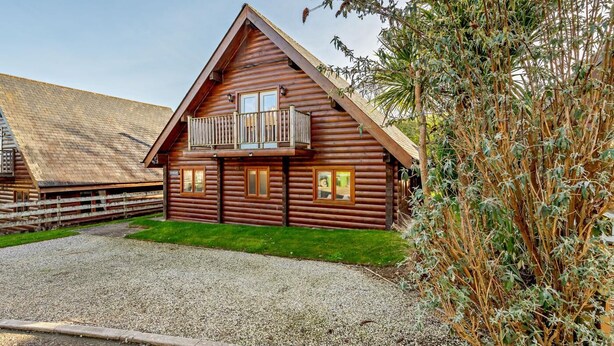A log cabin with a balcony, a gravel driveway, and a fenced area.