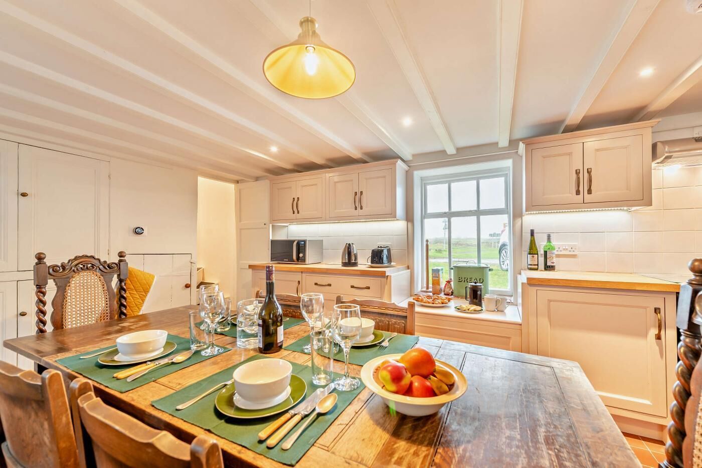 A well-lit kitchen with wooden cabinets, a dining table set with plates, glasses, and a bowl of fruit.