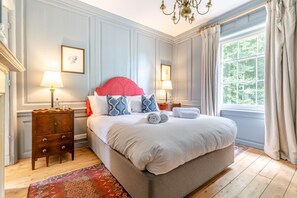 Un grand lit avec une tête de lit rouge, deux tables de chevet avec des lampes, une commode en bois et une vue sur la verdure par la fenêtre.