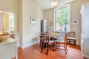 Un coin repas avec une table dressée pour quatre personnes, entouré de chaises en bois dotées d’assises rembourrées. On aperçoit un réfrigérateur au fond, et une fenêtre laisse entrer la lumière naturelle.