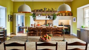 Een eetgedeelte met een houten tafel en stoelen, een schaal fruit en hangende keukengerei.