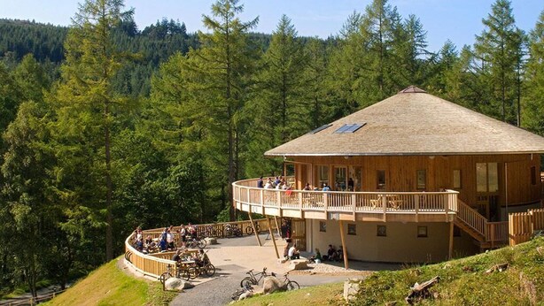 A wooden lodge with a balcony and a circular seating area, surrounded by a forested landscape. - 2 Bed in Maentwrog (oc-g28262) (Maentwrog)