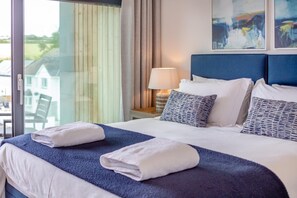 A hotel room with a large bed, two pillows, a nightstand with a lamp, and a view of a building through the window. - 2 Bed in Harlyn Bay (oc-a28321) (Harlyn Bay)