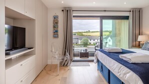 A modern hotel room with a flat-screen TV, a bed with a blue headboard, a clear chair, and a view of the outside. - 2 Bed in Harlyn Bay (oc-a28321) (Harlyn Bay)