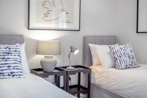 Une chambre d’hôtel avec deux lits, une table de chevet avec une lampe, une œuvre d’art encadrée au mur et une petite table de nuit avec un téléphone et un chargeur.