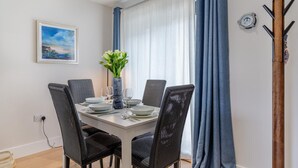 A dining area with a table set for four, surrounded by wicker chairs, a vase with flowers, and a painting on the wall. - 2 Bed in Harlyn Bay (oc-a28321) (Harlyn Bay)