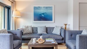 A modern living room with a grey sofa set, a coffee table with a plate of muffins, a framed picture of an underwater scene, and a floor lamp. - 2 Bed in Harlyn Bay (oc-a28321) (Harlyn Bay)