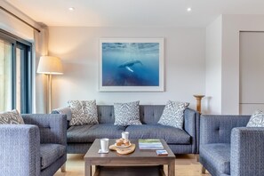 A modern living room with a grey sofa set, a coffee table with a plate of muffins, a framed picture of an underwater scene, and a floor lamp. - 2 Bed in Harlyn Bay (oc-a28321) (Harlyn Bay)