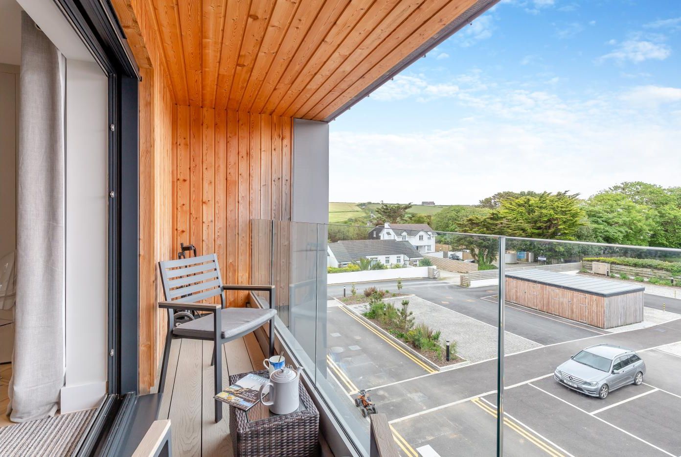 A balcony with a wooden ceiling, a wicker chair, a table with a teapot and a magazine, and a view of a parking area and houses in the background.
