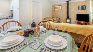 Ein Essbereich mit Tisch für vier Personen, eine Küche im Hintergrund, ein Wohnzimmer mit Sofa und Kamin sowie ein Fernseher auf einem Lowboard.