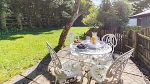 Eine Terrasse mit einem runden Tisch, Stühlen, einer Karaffe, Gläsern und einem Teller Essen.