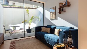 Un salon moderne avec un canapé bleu, un coin repas comprenant une table et des chaises, une grande plante en pot et une bouteille de vin posée sur une table d’appoint.