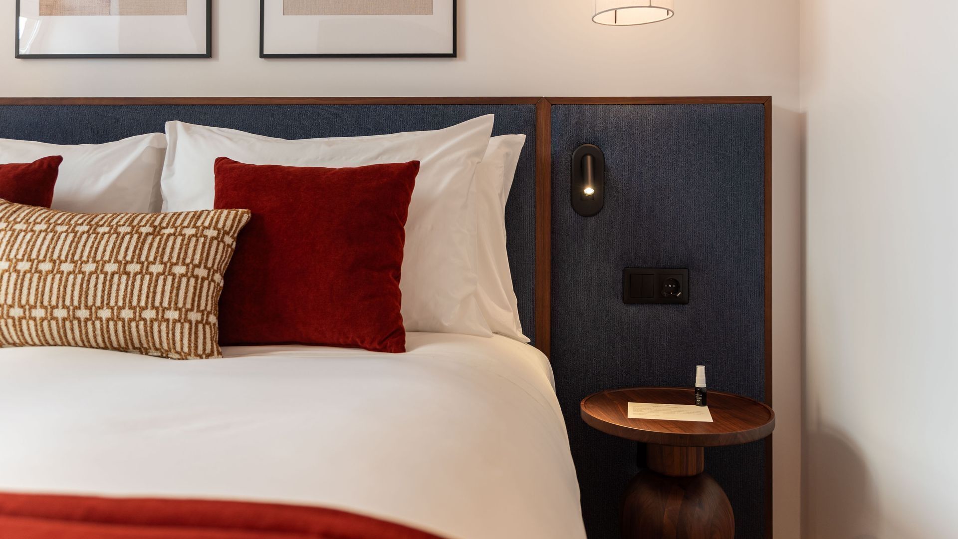 A neatly made bed with white and red pillows, a patterned pillow, and a small wooden side table with a lamp.