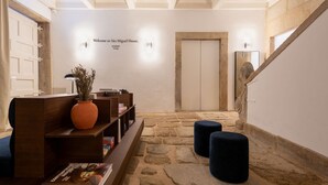 Un área de recepción con suelo de piedra, un cartel de bienvenida, mostrador de madera, una planta en maceta y una escalera.