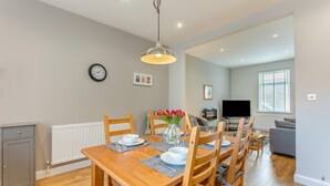 A dining area with a wooden table, chairs, and a vase of flowers. There is a clock on the wall and a television in the background. - 2 Bed in Amble (oc-b28345) (Amble)