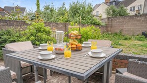 Uma mesa no pátio com cadeiras de vime, uma cesta de frutas e itens de café da manhã.