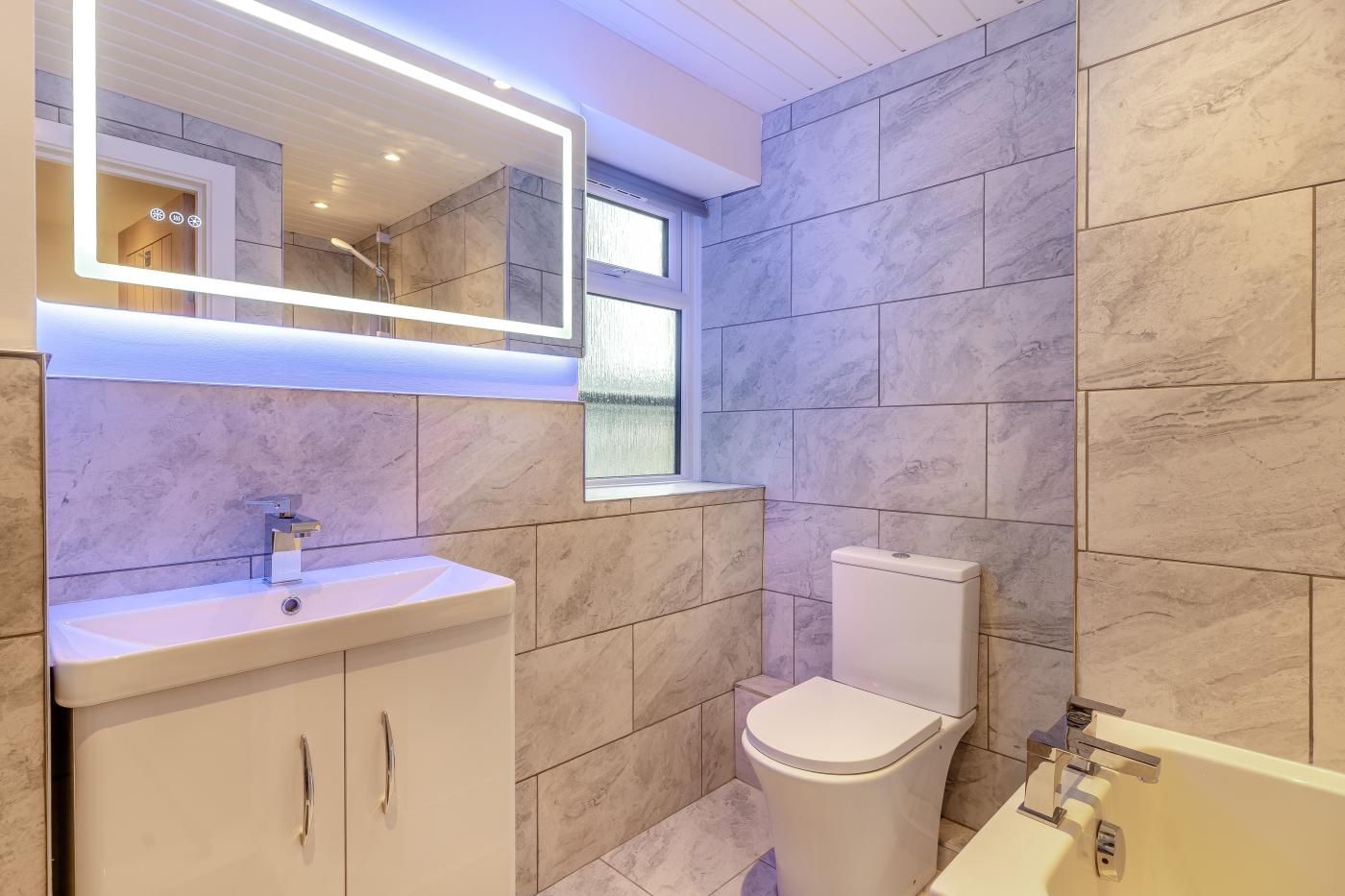 A modern bathroom with a white sink, mirror, and toilet, and a window above the toilet.