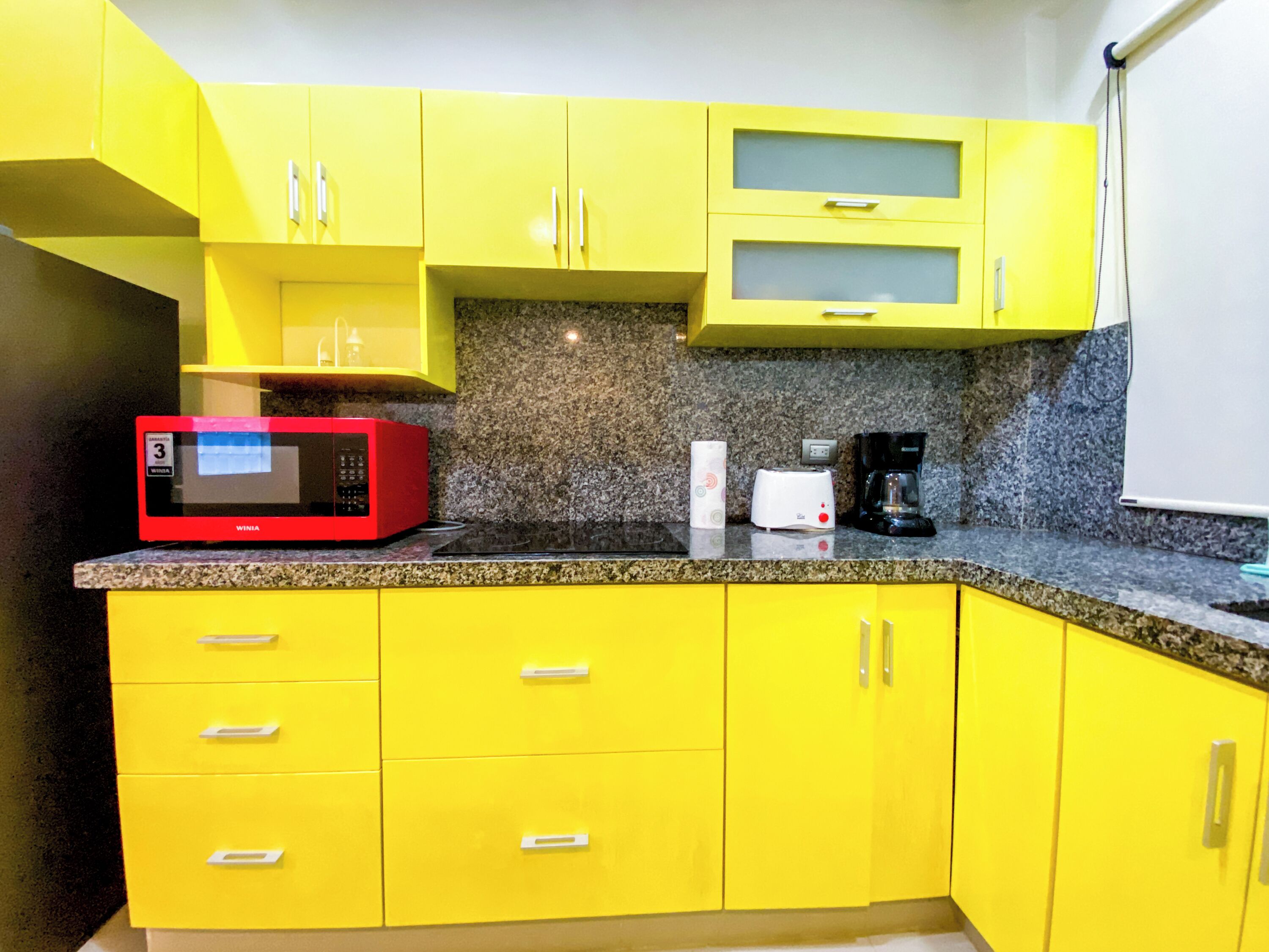 Une cuisine avec des meubles de couleur jaune, un four à micro-ondes rouge, un grille-pain et une machine à café, le tout posé sur un plan de travail en granit.