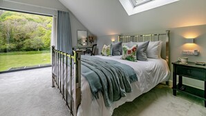 Ein Schlafzimmer mit Oberlicht, einem Bett mit Blumenkissen, einem Nachttisch mit Lampe und Blick auf einen grünen Garten.