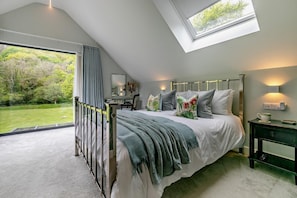 A bedroom with a skylight, a bed with floral pillows, a bedside table with a lamp, and a view of a green garden.