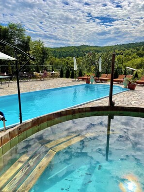 Une piscine aux eaux cristallines, dotée d’une terrasse en bois, entourée de chaises longues et de parasols, le tout se fondant dans un décor luxuriant de verdure et de collines.