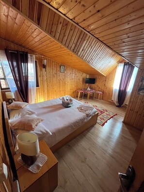 Une chambre dans un chalet en bois, avec un lit, une petite table et une lampe, un téléviseur et une fenêtre avec des rideaux.