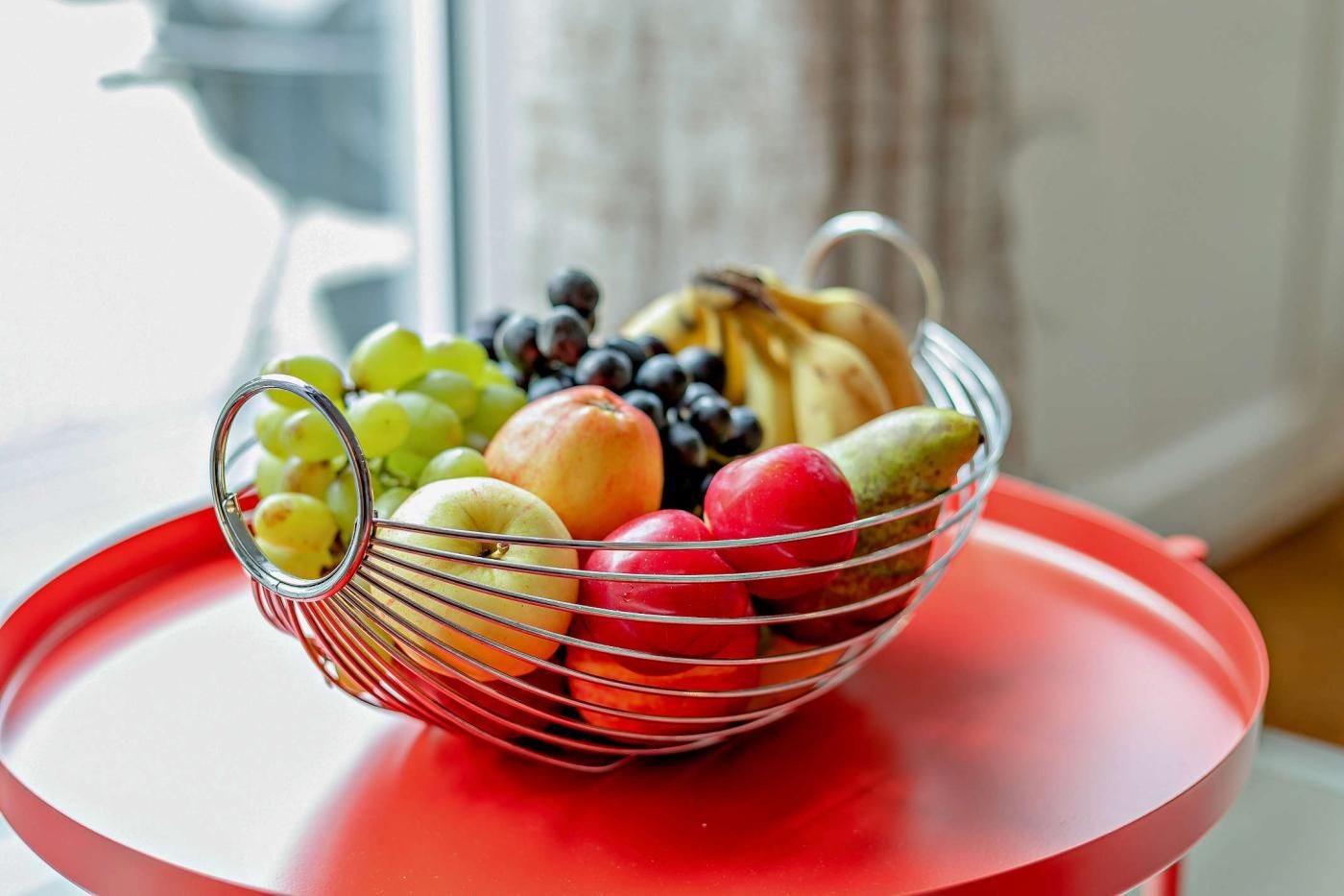 Un panier de fruits est posé sur une table rouge, avec une fenêtre en arrière-plan.