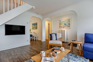 A modern living room with a flat-screen TV, a dining area with a table and chairs, a blue sofa, and a staircase. - 3 Bed in Porthleven (oc-s28377) (Porthleven)
