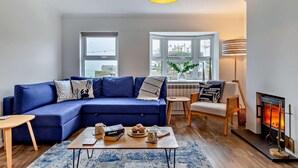 Ein modernes Wohnzimmer mit einem blauen Sofa, einem kleinen Tisch mit Büchern und einer Tasse, einem Kamin und großen Fenstern mit Stadtblick.