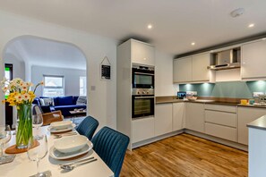 A modern kitchen with white cabinets, a built-in oven, and a dining area with a table set for two. - 3 Bed in Porthleven (oc-s28377) (Porthleven)