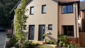 Un immeuble résidentiel moderne à deux étages, doté d’un toit plat, de grandes fenêtres et d’un jardin bien entretenu à l’avant, avec un petit arbre et des parterres de fleurs.