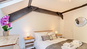 A bedroom with a sloped ceiling, a bed with white bedding, two bedside tables with lamps, a potted orchid, and a round mirror on the wall. - 2 Bed in Tenby (oc-f28369) (Tenby)