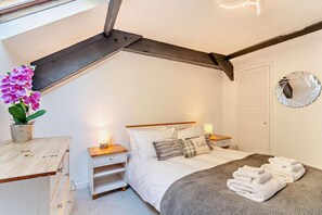Ein Schlafzimmer mit schrägen Wänden, einem Bett mit weißer Bettwäsche, zwei Nachttischen mit Lampen, einer Topforchidee und einem runden Spiegel an der Wand.