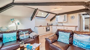 A cozy living room with leather sofas, a wooden coffee table, and decorative pillows. A kitchen with white cabinets and a clock is visible in the background. - 2 Bed in Tenby (oc-f28369) (Tenby)