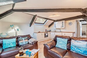 Ein gemütliches Wohnzimmer mit Ledersofas, einem Holztisch und dekorativen Kissen. Im Hintergrund ist eine Küche mit weißen Schränken und einer Uhr zu sehen.