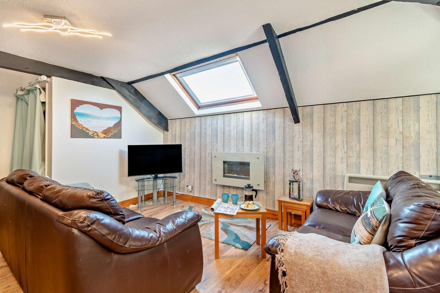 A living room with a sloped ceiling, a flat-screen TV, leather sofas, a coffee table, and a small table with a kettle and cups.