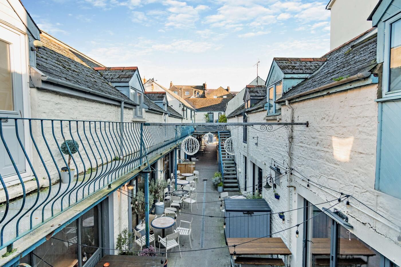 A narrow alleyway with buildings on either side, outdoor seating, and a metal railing.