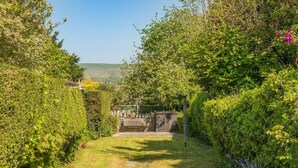 Ein Gartenweg führt zu einem hölzernen Tor, umgeben von dichtem Grün und blauem Himmel.