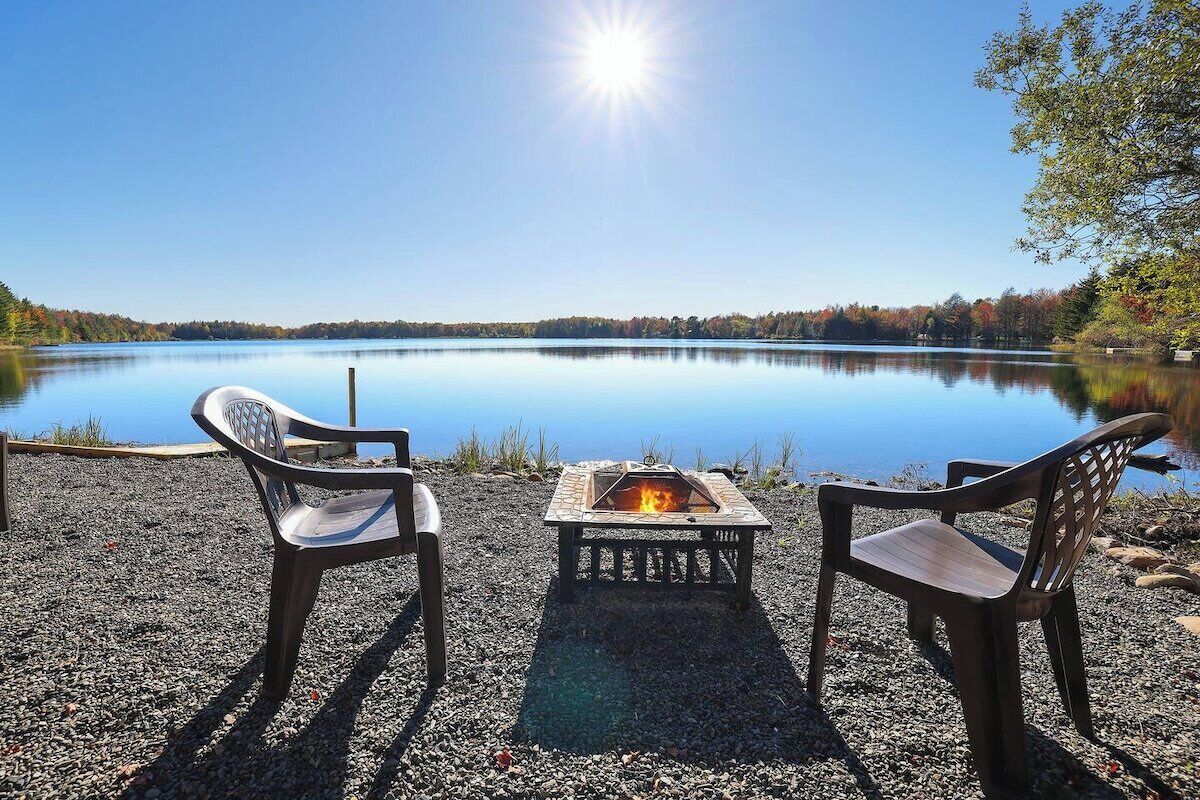 A lakeside setting with two chairs facing a fire pit, surrounded by pebbles and a clear view of the lake and trees.