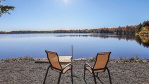 A serene lakeside setting with two chairs and a wooden table facing the water, surrounded by pebbles and autumn foliage. - Backyard lake-Boat– Kayaks • Firepit • Game room• pool access • Pet-friendly (Coolbaugh Township)