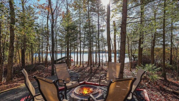 A fire pit with four chairs arranged around it, set in a forested area with a lake in the background. - Backyard lake-Boat– Kayaks • Firepit • Game room• pool access • Pet-friendly (Coolbaugh Township)