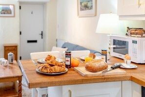 Une table de petit-déjeuner avec des croissants, du pain, de la confiture et du jus d’orange, posée sur une table en bois dans une cuisine aux armoires blanches et équipée d’un four à micro-ondes.