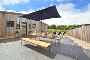 An outdoor patio with a wooden table and benches, a large umbrella, and blue lounge chairs. - 10 Bed in Briston (oc-mjbrdb) (Briston)