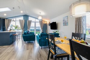 A modern kitchen with a central island, a dining area with a wooden table and chairs, and a living area with blue sofas. - 2 Bed in Burnham Market (oc-n28359) (Burnham Market)