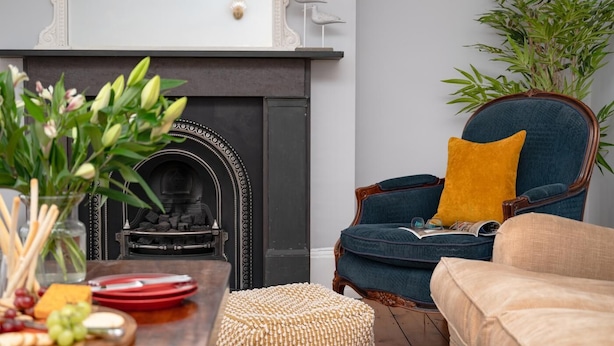 Una sala de estar con chimenea, un sillón azul con cojín mostaza, un reposapiés de mimbre y una mesa con aperitivos.