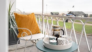 Un balcón con barandilla de metal blanco, una silla blanca con cojín color mostaza, y una pequeña mesa redonda con una bandeja que contiene una cafetera, tazas y un platillo.