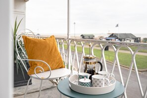 A balcony with a white metal railing, a white chair with a mustard-colored cushion, a small round table with a tray holding a coffee pot, cups, and a saucer. - 4 Bed in Aldeburgh (oc-e28371) (Aldeburgh)