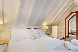 A bedroom with a sloped ceiling, a bed with white linens, a nightstand with a lamp, a dresser, and a door leading to another room. - 2 Bed in Bodham (oc-m15008) (Bodham)