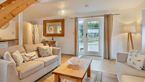 A living room with a sofa, coffee table, and a dining area with a table and chairs. There is a large window with curtains, a wooden ceiling beam, and a framed picture on the wall. - 2 Bed in Bodham (oc-m15008) (Bodham)